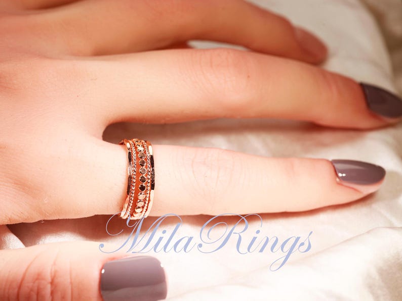 Puede incluir: Un anillo en tono oro rosa con una banda central de peque&ntilde;as piedras circulares oscuras, flanqueada por dos filas de detalles met&aacute;licos texturizados y con cuentas. El anillo tiene un perfil ancho y plano. El texto Mila Rings es visible.