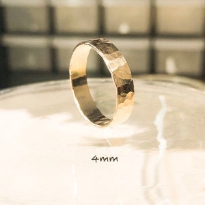 K&ouml;nnte beinhalten: Ein goldener, geh&auml;mmerter Bandring mit einer Breite von 4 mm. Der Ring liegt auf einer durchsichtigen Oberfl&auml;che.