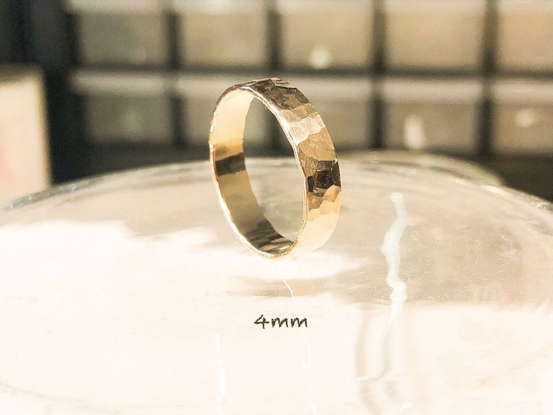 K&ouml;nnte beinhalten: Ein goldener, geh&auml;mmerter Bandring mit einer Breite von 4 mm. Der Ring liegt auf einer durchsichtigen Oberfl&auml;che.