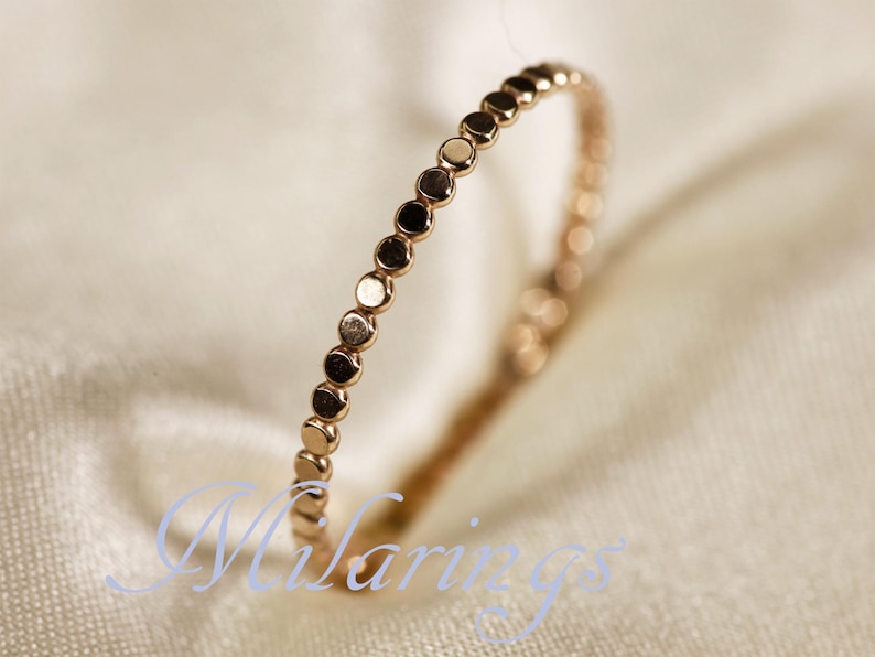 May include: A delicate rose gold ring featuring a series of small, evenly spaced circular accents. The ring is set against a soft, off-white background, highlighting its polished finish and minimalist design. The ring's design is simple and elegant.