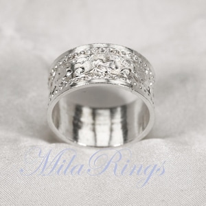 May include: A silver ring with an embossed design. The ring has a wide band with a detailed pattern of floral and scrollwork. The ring is displayed on a white, textured surface. The ring is a piece of jewellery.