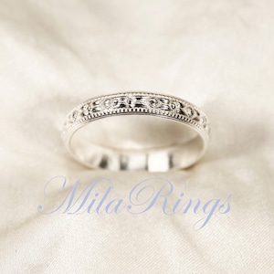May include: A silver wedding band with a delicate, intricate design. The band is smooth and polished, with a subtle sheen.