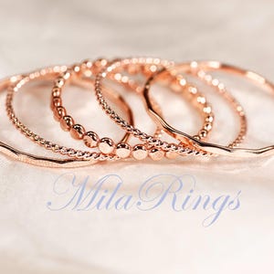 May include: A set of five rose gold rings with varying textures, including smooth, twisted, and beaded designs. The rings are arranged in a cluster, and the text "Mila Rings" is printed in a light blue script font across the lower portion of the image.