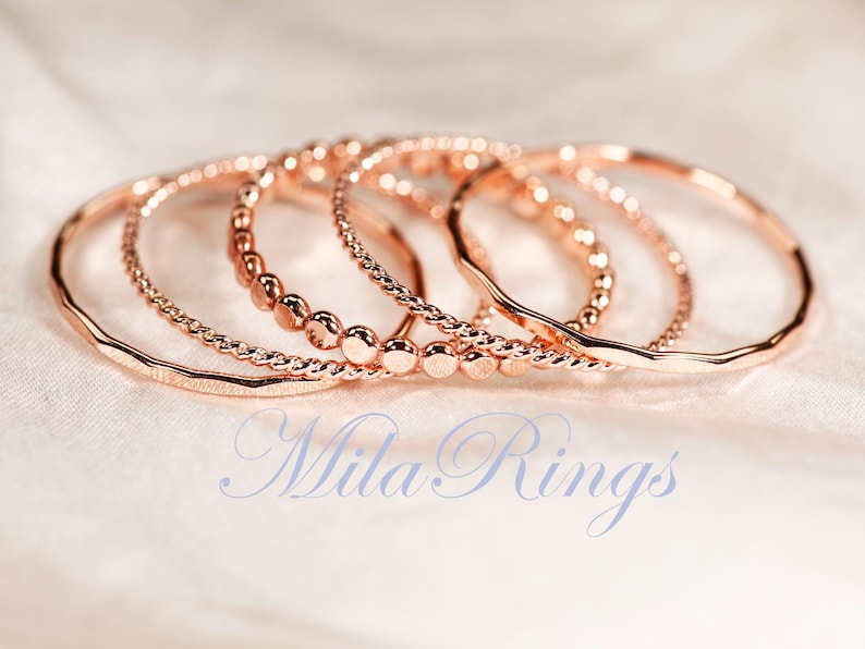 Puede incluir: Un conjunto de cinco anillos de oro rosa con texturas variadas, incluyendo dise&ntilde;os lisos, trenzados y con cuentas. Los anillos est&aacute;n dispuestos en grupo y el texto "Mila Rings" est&aacute; impreso en una fuente cursiva azul claro en la parte inferior.