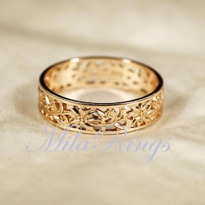 May include: A gold-colored ring with an intricate floral and scrollwork design. The ring is a band style, with the decorative pattern extending around the entire circumference. The ring is set against a soft, neutral background.