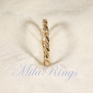 Peut inclure: Une bague dor&eacute;e avec un motif tress&eacute;. La bague est pos&eacute;e sur un fond blanc cass&eacute; et doux. Le texte "Mila Rings" est visible dans la partie inf&eacute;rieure de l'image.