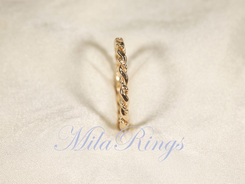 Peut inclure: Une bague dor&eacute;e avec un motif tress&eacute;. La bague est pos&eacute;e sur un fond blanc cass&eacute; et doux. Le texte "Mila Rings" est visible dans la partie inf&eacute;rieure de l'image.