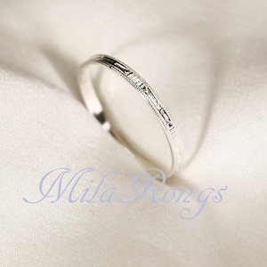 May include: A silver ring with an engraved design. The ring is set against a white, textured background. The ring's design features a series of small, repeating patterns. The text "Mila Rings" is visible in the image.