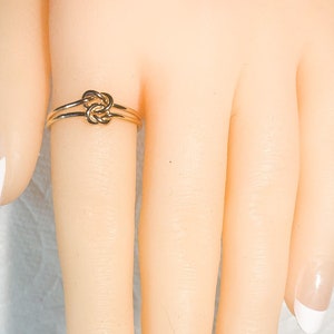 May include: A gold knot ring with a simple design. The ring is worn on a model's finger.