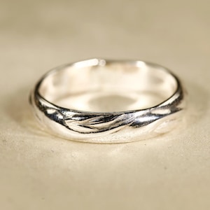 May include: A silver ring with a textured, organic design. The ring has a smooth interior and a wavy, textured exterior. The ring is displayed on a neutral-toned surface, highlighting its metallic sheen. A piece of jewelry.