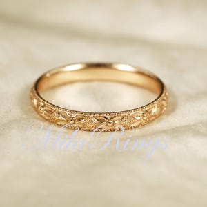 May include: A gold-colored ring with an intricate floral pattern around the band. The ring is set against a soft, off-white background. The ring's design features small, repeating floral and leaf motifs. The text "Atelier Rings" is visible in the image.