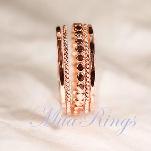 Puede incluir: Un conjunto de cuatro anillos de oro rosa apilados, con texturas variadas que incluyen una banda martillada, una banda de cuerda trenzada y una banda con peque&ntilde;os discos circulares. El texto Mila Rings es visible en azul en la parte inferior.
