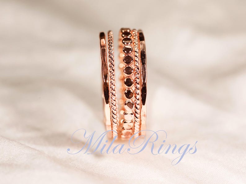Puede incluir: Un conjunto de cuatro anillos de oro rosa apilados, con texturas variadas que incluyen una banda martillada, una banda de cuerda trenzada y una banda con peque&ntilde;os discos circulares. El texto Mila Rings es visible en azul en la parte inferior.