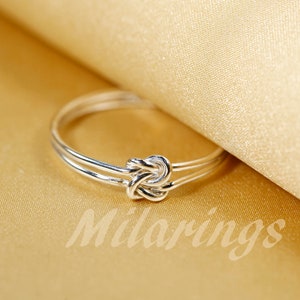 May include: A silver ring with a knot design. The ring is on a gold fabric background.