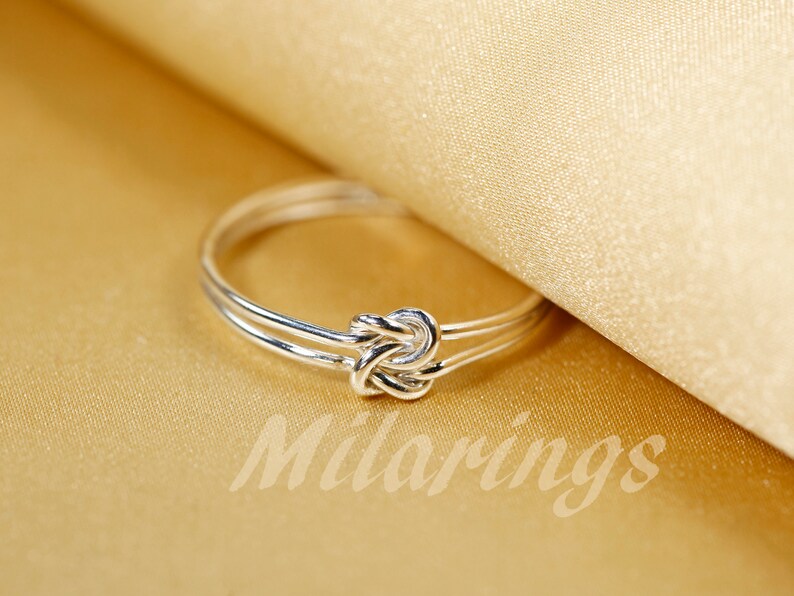 May include: A silver ring with a knot design. The ring is on a gold fabric background.