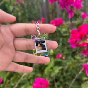 May include: A rectangular clear resin keychain featuring a small embedded photograph of a golden-colored dog. The resin is decorated with tiny dried flowers in shades of green, blue, and yellow, and includes a silver-toned metal chain and ring.