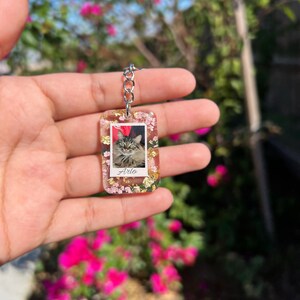 May include: A rectangular resin keychain featuring a central photograph of a fluffy cat with the name Arlo printed below. The clear resin is embedded with small pink dried flowers and gold leaf flakes, attached to a silver-toned metal chain link.