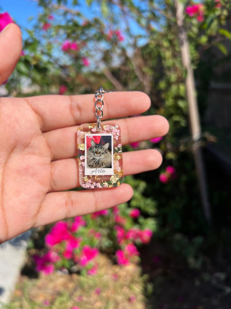 May include: A rectangular resin keychain featuring a central photograph of a fluffy cat with the name Arlo printed below. The clear resin is embedded with small pink dried flowers and gold leaf flakes, attached to a silver-toned metal chain link.