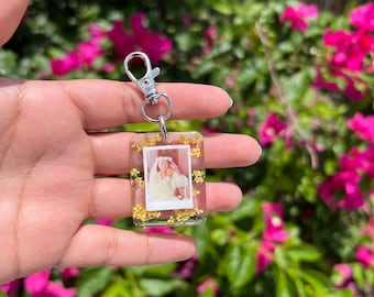 May include: A rectangular clear resin keychain featuring a small embedded photograph of a person wearing a white veil and dress, surrounded by tiny yellow dried flowers, attached to a silver-toned metal lobster claw clasp and jump ring.