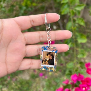 May include: A rectangular resin keychain featuring an embedded photograph of a person holding a cat. The clear resin is decorated with small blue dried flowers and gold leaf flakes, attached to a silver-toned metal split ring and chain.