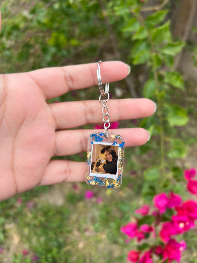 May include: A rectangular resin keychain featuring an embedded photograph of a person holding a cat. The clear resin is decorated with small blue dried flowers and gold leaf flakes, attached to a silver-toned metal split ring and chain.