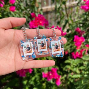 May include: Three rectangular clear resin keychains featuring embedded miniature photographs of a young child wearing a black t-shirt. The resin is decorated with silver and blue metallic flakes, and each keychain is attached to a silver metal chain.