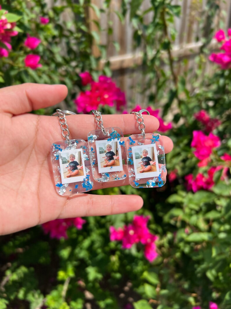 May include: Three rectangular clear resin keychains featuring embedded miniature photographs of a young child wearing a black t-shirt. The resin is decorated with silver and blue metallic flakes, and each keychain is attached to a silver metal chain.