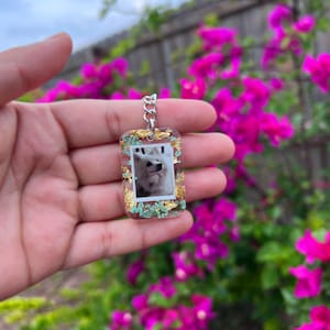May include: A rectangular clear resin keychain featuring an embedded photograph of a white dog. The keychain is decorated with gold leaf flakes and small light blue dried flowers, attached to a silver-toned metal jump ring and chain link.