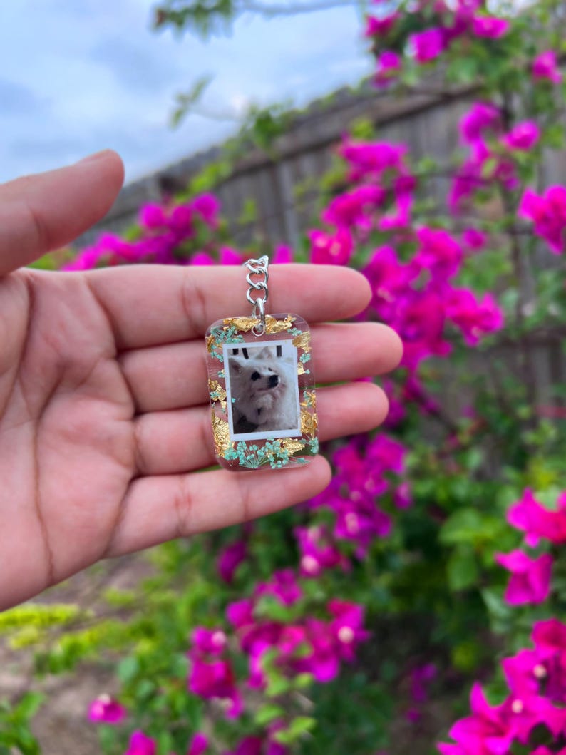 May include: A rectangular clear resin keychain featuring an embedded photograph of a white dog. The keychain is decorated with gold leaf flakes and small light blue dried flowers, attached to a silver-toned metal jump ring and chain link.