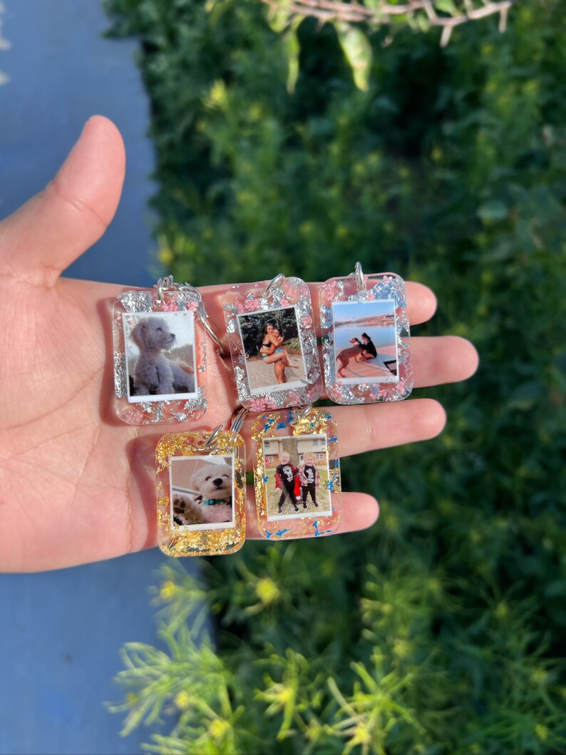 May include: Five rectangular resin keychains featuring embedded photographs and metallic flakes. Three keychains contain silver flakes with pink accents, while two feature gold flakes. Each keychain displays a unique personal photo.