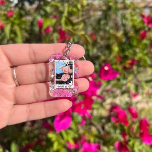 May include: A rectangular clear resin keychain featuring embedded pink dried flowers and silver foil flakes. The center contains a small photograph of two people with the text "love u <3" printed in black ink at the bottom of the photo insert.