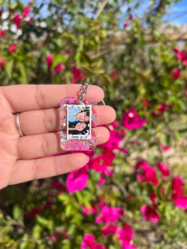May include: A rectangular clear resin keychain featuring embedded pink dried flowers and silver foil flakes. The center contains a small photograph of two people with the text "love u <3" printed in black ink at the bottom of the photo insert.