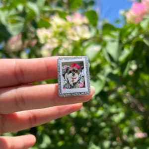 May include: A rectangular pin featuring a miniature photograph of a small dog with a pink bow and collar. The image is framed by a border covered in small, clear, reflective crystals that catch the light, creating a sparkling effect.