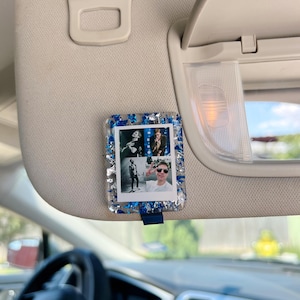May include: A rectangular resin car visor clip featuring a collage of four small photographs of a person. The clear resin frame is embedded with metallic blue and silver foil flakes, creating a decorative border around the central photo prints.