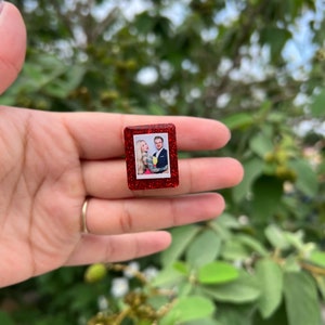 May include: A red glitter picture frame with a photo of a couple inside. The frame is small and is being held in a person's hand.