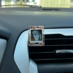 May include: A rectangular car air freshener clip featuring a central square photograph of a small white puppy. The frame surrounding the photo is clear with embedded gold leaf flakes and small pink dried flowers, attached to a car air vent.
