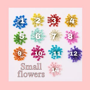 May include: A collection of thirteen small dried flowers arranged in a grid, each labeled with a white number from 1 to 12 and 14. The flowers are dyed in various colors including blue, yellow, red, green, turquoise, white, pink, orange, and purple.
