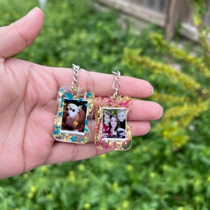 May include: Two rectangular resin keychains featuring embedded photographs. One keychain contains a photo of a small dog and blue floral accents, while the other holds a photo of three people with pink floral accents. Both include gold leaf flakes.