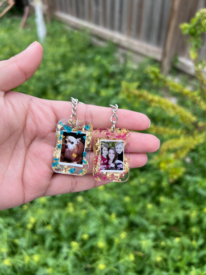 May include: Two rectangular resin keychains featuring embedded photographs. One keychain contains a photo of a small dog and blue floral accents, while the other holds a photo of three people with pink floral accents. Both include gold leaf flakes.