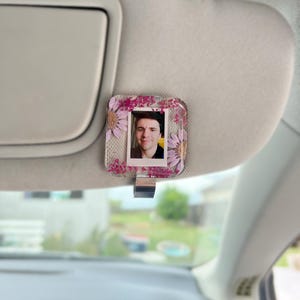 May include: A square resin car visor clip featuring a central portrait photograph surrounded by embedded pink and purple dried flowers. The clear resin frame has a metallic silver clip attached to the bottom edge for securing onto a vehicle sun visor.