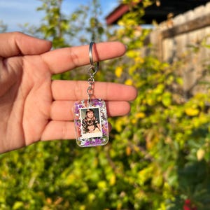 May include: A rectangular clear resin keychain featuring a small embedded photograph of two people. The resin is filled with purple and white dried flowers and silver flakes. The keychain is attached to a silver metal split ring and chain.