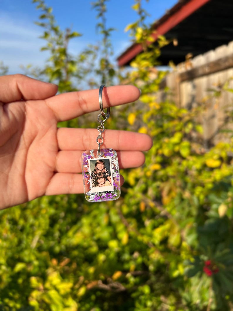 May include: A rectangular clear resin keychain featuring a small embedded photograph of two people. The resin is filled with purple and white dried flowers and silver flakes. The keychain is attached to a silver metal split ring and chain.