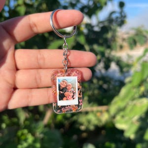 May include: A rectangular clear resin keychain featuring an embedded miniature photograph of two people. The resin is decorated with small pink and orange dried flowers and gold leaf flakes. It is attached to a silver metal split ring by a chain.