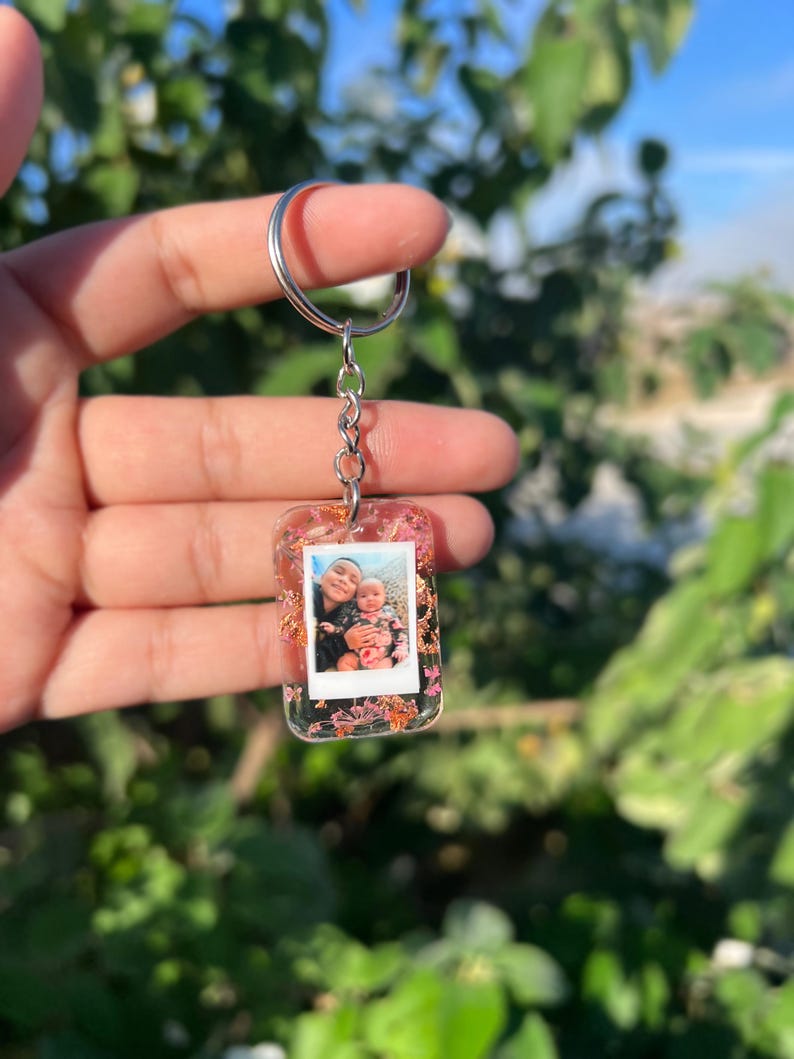 May include: A rectangular clear resin keychain featuring an embedded miniature photograph of two people. The resin is decorated with small pink and orange dried flowers and gold leaf flakes. It is attached to a silver metal split ring by a chain.