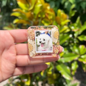 May include: A square resin charm featuring a central photograph of a white dog with its tongue out. The clear resin is embedded with dried white flowers and gold leaf flakes, creating a decorative border around the small rectangular photo.