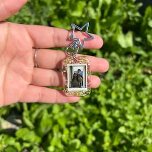 May include: A silver star-shaped keyring with a clear rectangular charm containing a photo of a couple. The charm is filled with gold glitter and small white flowers.