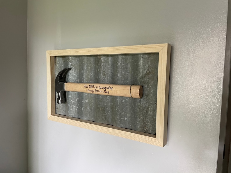 May include: A framed wood and metal sign with a hammer inside. The hammer has a wooden handle with the text "Our Dad can fix anything Happy Father's Day" engraved on it.