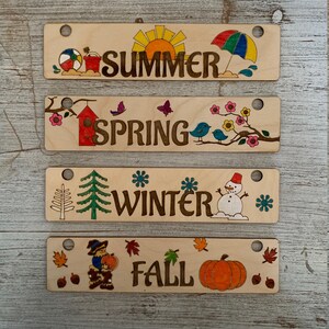 Pode incluir: Quatro placas de madeira com imagens e texto que representam as quatro esta&ccedil;&otilde;es: ver&atilde;o, primavera, inverno e outono. Cada placa tem um furo na parte superior para pendurar.