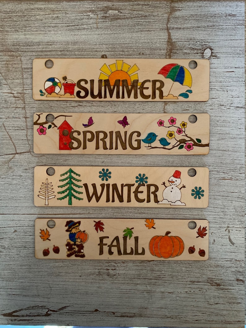 Pode incluir: Quatro placas de madeira com imagens e texto que representam as quatro esta&ccedil;&otilde;es: ver&atilde;o, primavera, inverno e outono. Cada placa tem um furo na parte superior para pendurar.