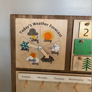 Pode incluir: Um quadro de previs&atilde;o do tempo de madeira com um dial rotativo que apresenta diferentes &iacute;cones meteorol&oacute;gicos: ensolarado, nublado, chuvoso, nevado, ventoso e parcialmente ensolarado. O quadro tamb&eacute;m inclui um calend&aacute;rio com os dias da semana e os dias numerados.